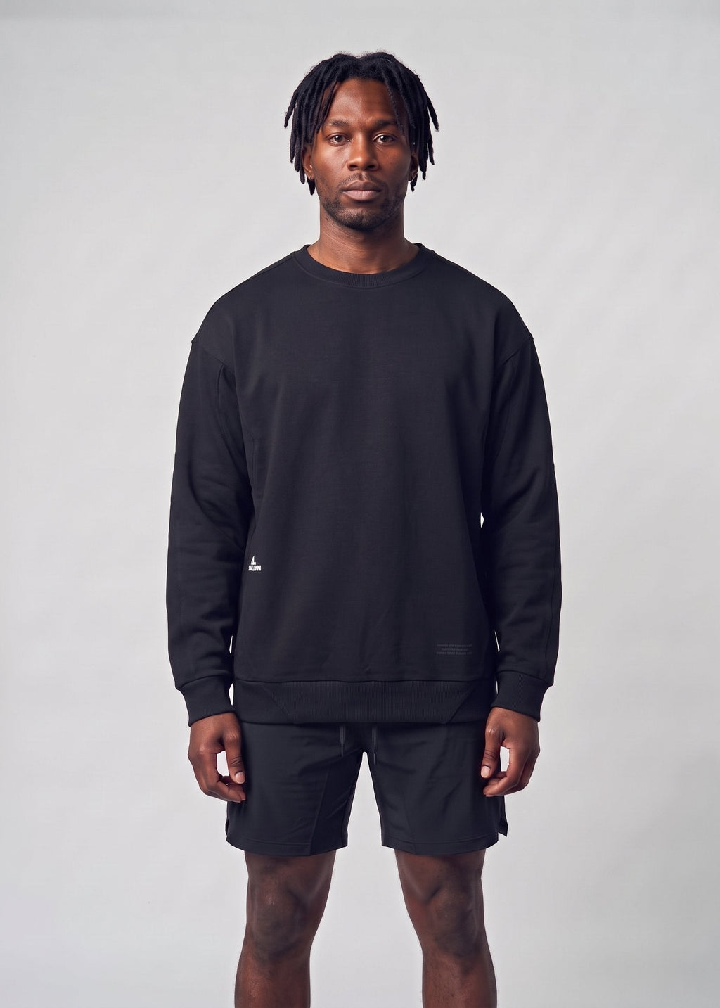 A man modeling a black crewneck sweatshirt and matching black shorts against a light gray studio background.