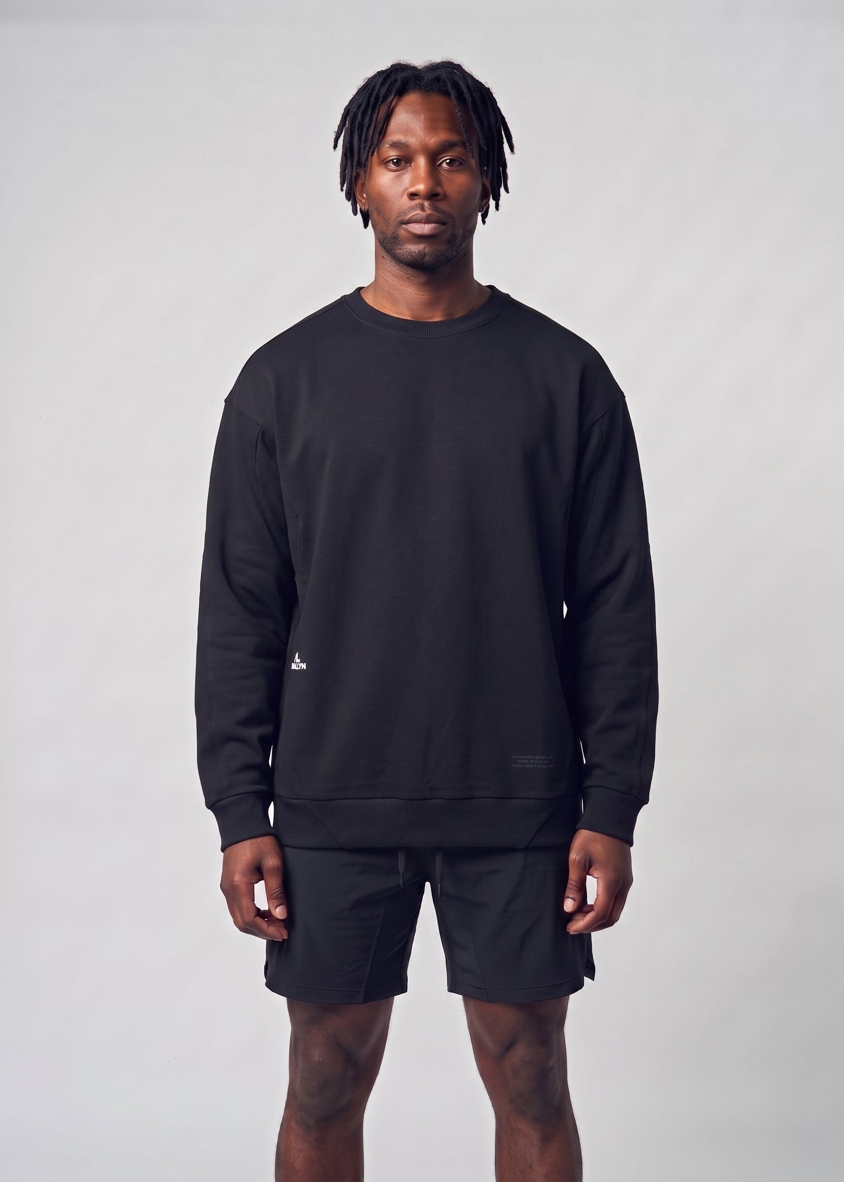 A man modeling a black crewneck sweatshirt and matching black shorts against a light gray studio background.