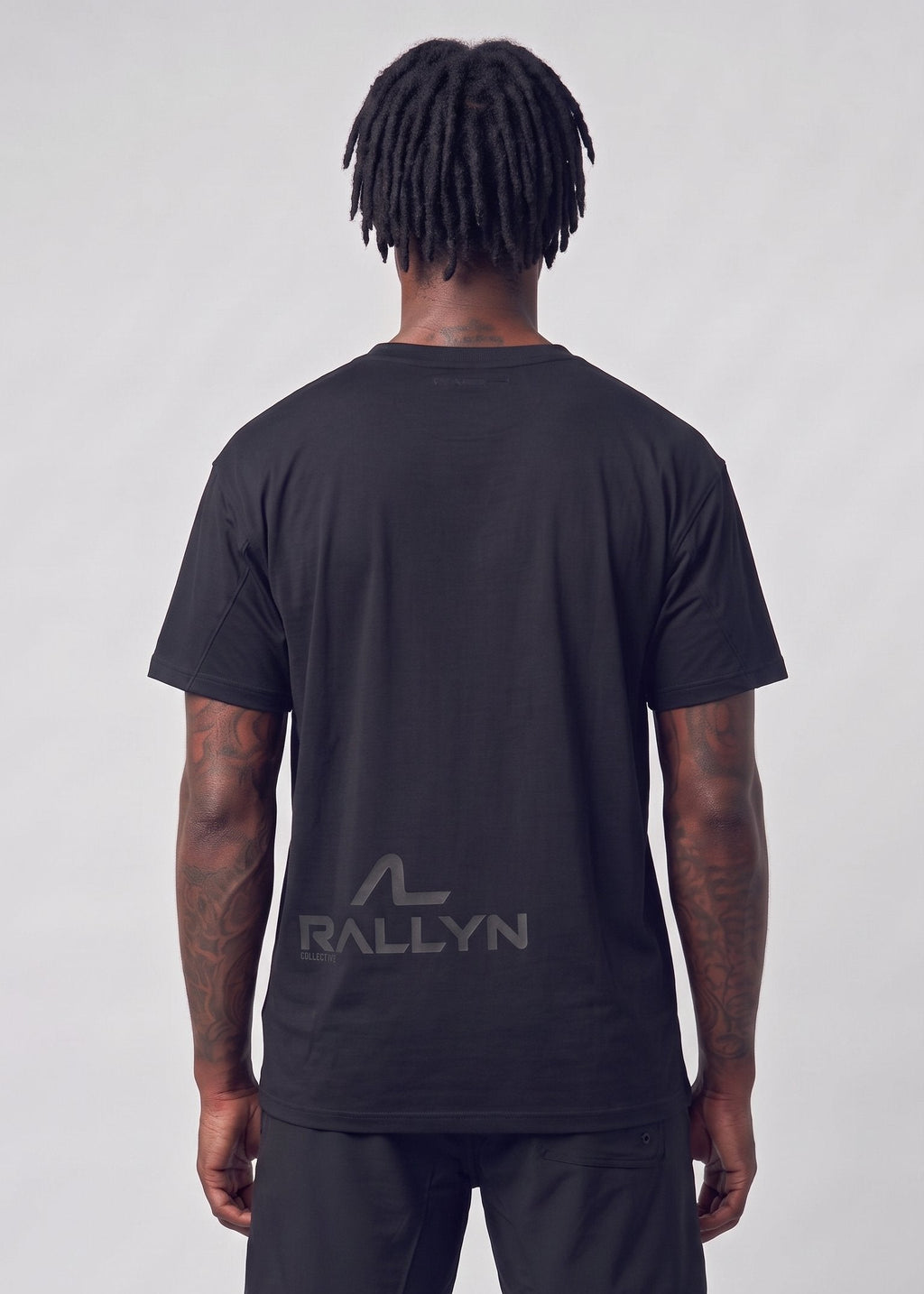 A man viewed from behind wearing a black short-sleeve t-shirt and black shorts, featuring a dark gray 'RALLYN' logo printed on the back of the shirt, against a light gray studio background.
