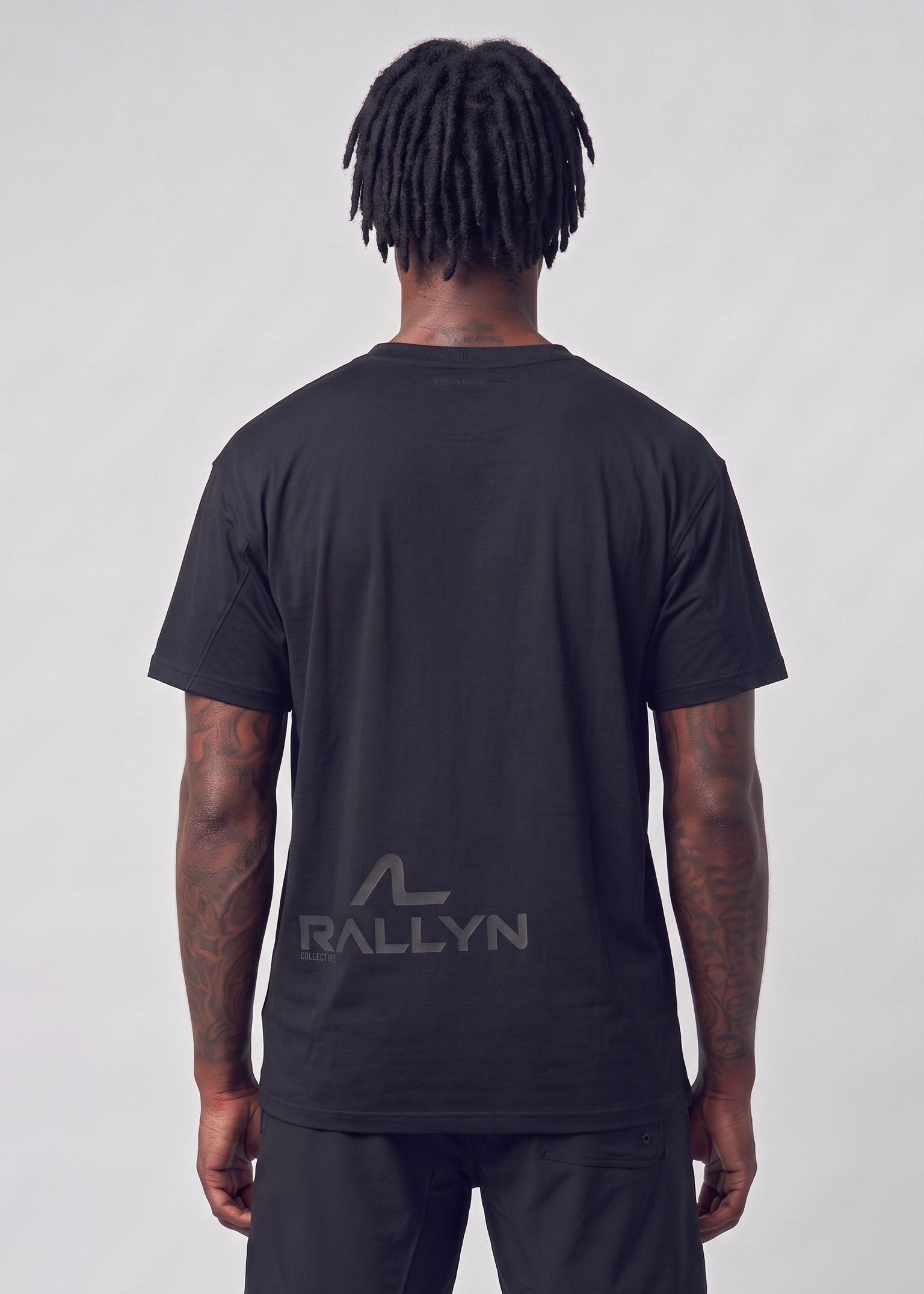 A man viewed from behind wearing a black short-sleeve t-shirt and black shorts, featuring a dark gray 'RALLYN' logo printed on the back of the shirt, against a light gray studio background.