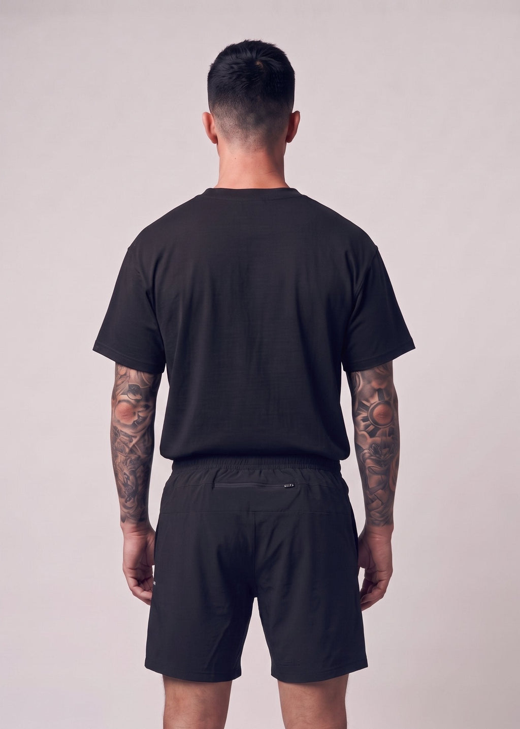 A man viewed from behind wearing a plain black short-sleeve t-shirt and black athletic shorts against a light gray studio background.