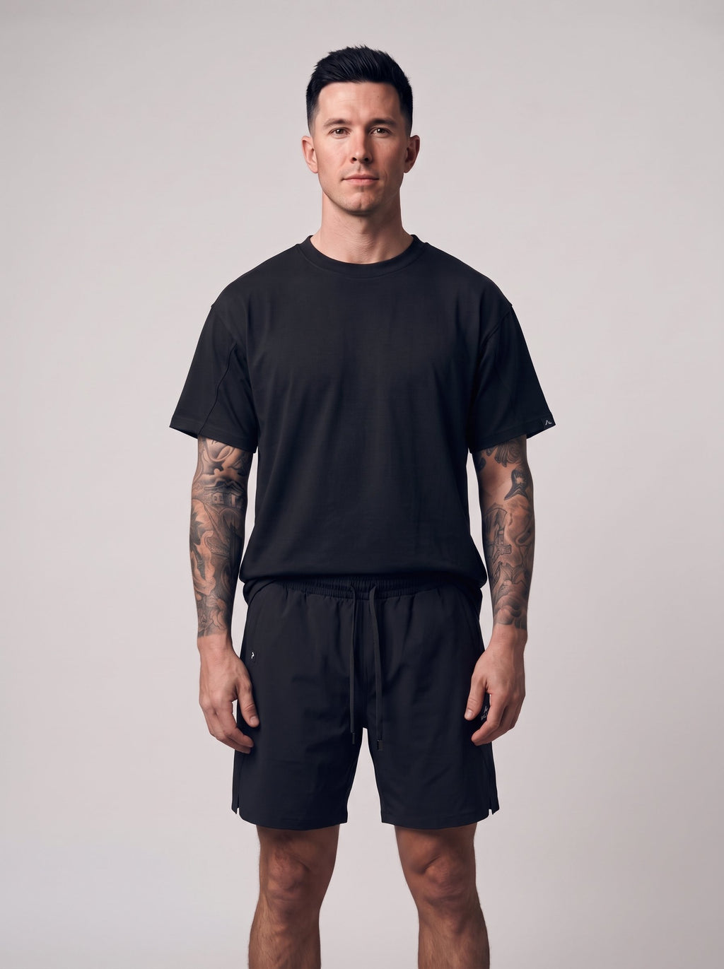 A man with tattoos stands against a light gray studio background wearing a plain black short-sleeve crewneck t-shirt and matching black shorts.